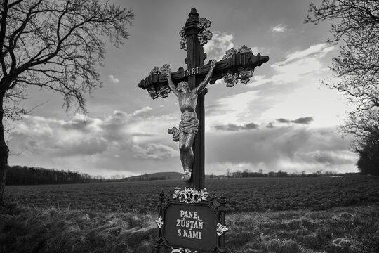 "Lord stay with us" is written on a Christ cross in Czech language. The cross stands in front of a green field. In the background are clouds and the sun. INRI.