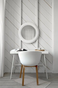White chair at table with book and cup of beverage placed near wall with round mirror in light room designed in minimal style