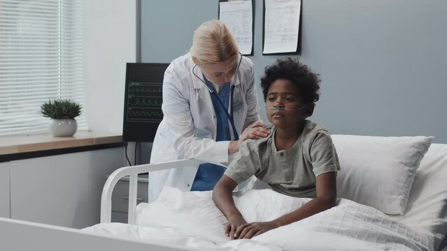 Medium long of Black boy with oxygen tube sitting on bed in hospital room at daytime, mature Caucasian female doctor using stethoscope on his back, talking to child