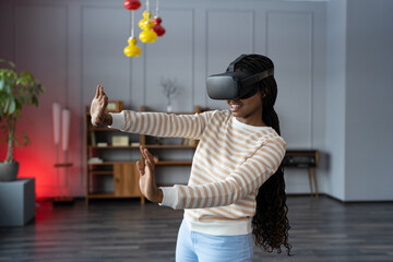 Young amazed astonished african woman testing VR goggles touching objects with hands in virtual reality at home. Excited black millennial girl using augmented reality at first time