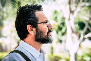 Side portrait of caucasian adult mature man with beard and eyewear looking in front with park nature geen defocused background. Serious and professional male people in outdoor