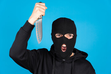 Angry assasin wearing black ski mask and holding and menacing with knife isolated on blue...