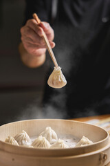 Close up of Chinese steamed Xiaolongbao held in chopsticks, being taken out of bamboo steamer with rustic wood.