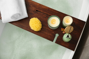 Wooden bath tray with candles, air freshener, towels and sponge on tub, top view