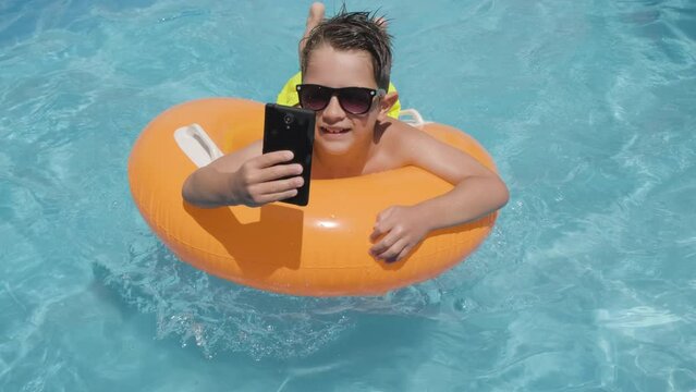 Young boy in Sunglasses using Cellphone on orange floater while relaxing in the swimming pool