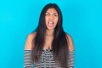 Fototapeta premium Body language. Disgusted stressed out young latin woman wearing striped T-shirt over blue background , frowning face, demonstrating aversion to something.