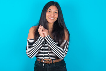 Dreamy charming young latin woman wearing striped T-shirt over blue background with pleasant expression, keeps hands crossed near face, excited about something pleasant.