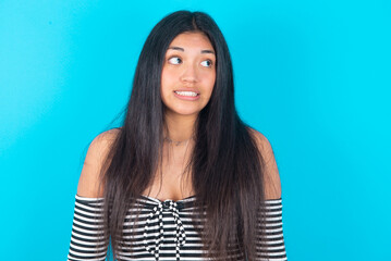 Fototapeta premium Oops! Portrait of young latin woman wearing striped T-shirt over blue background clenches teeth and looks confusedly aside, realizes her bad mistake,