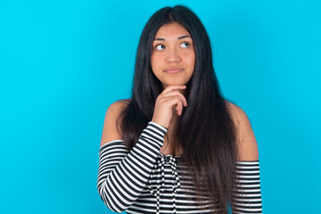 Dreamy young latin woman wearing striped T-shirt over blue background with pleasant expression, looks sideways, keeps hand under chin, thinks about something pleasant.
