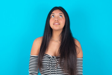Fototapeta premium young latin woman wearing striped T-shirt over blue background looking up as he sees something strange.