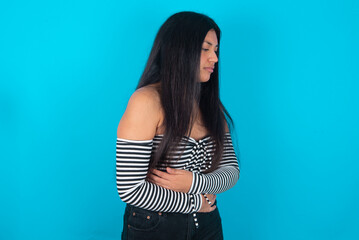 young latin woman wearing striped T-shirt over blue background suffering from strong stomachache.