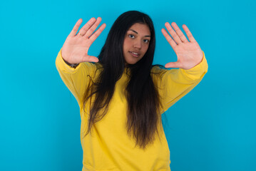 Portrait of smiling young latin woman wearing yellow sweater over blue background looking at camera and gesturing finger frame. Creativity and photography concept.