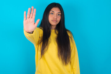 young latin woman wearing yellow sweater over blue background doing stop gesture with palm of the hand. Warning expression with negative and serious gesture on the face.