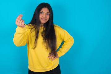 Fototapeta premium What the hell are you talking about. Shot of frustrated young latin woman wearing yellow sweater over blue gesturing with raised hand doing Italian gesture, frowning, being displeased and confused 