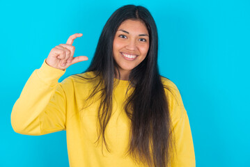 young latin woman wearing yellow sweater over blue background smiling and gesturing with hand small size, measure symbol.