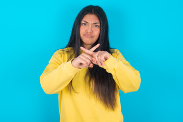 young latin woman wearing yellow sweater over blue background has rejection angry expression...