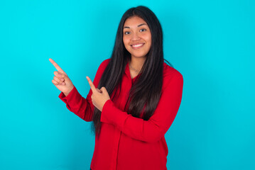 young latin woman wearing red shirt over blue background points at copy space indicates for...