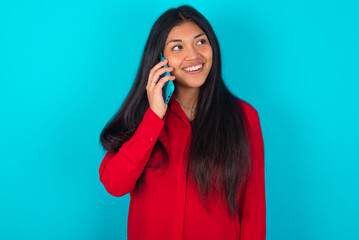 Pleasant looking happy young latin woman wearing red shirt over blue background has nice telephone conversation and looks aside, has nice mood and smiles positively while talks via cell phone