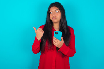 young latin woman wearing red shirt over blue background points thumb away and shows blank space aside, holds mobile phone for sending text messages.