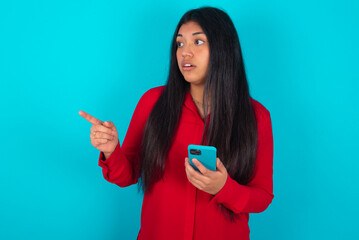 Stunned young latin woman wearing red shirt over blue background points sideways right copy space, recommends product, sees astonishing thing
