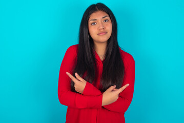 Obraz premium Serious young latin woman wearing red shirt over blue background crosses hands and points at different sides hesitates between two items. Hard decision concept