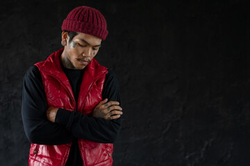Portrait of cool Asian hip hop young bearded man with tattoo on face, neck and hands standing and cross his arms with nose piercing, big ear piercing and wear red knit hat and vest. Black background