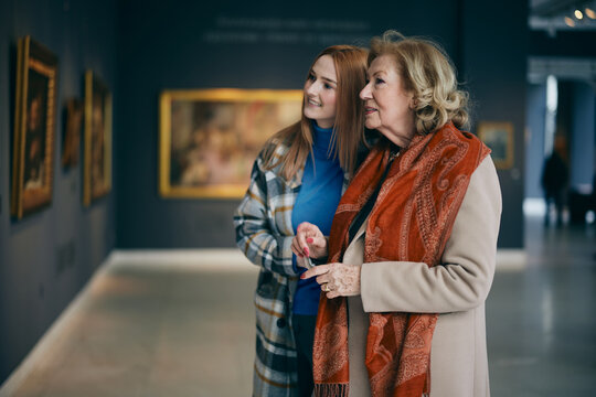 Grandmother and adolescent granddaughter are looking at the paintings in the art gallery.