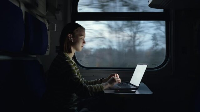 Young business woman in wireless headphones works while traveling by train, sits in a compartment at a table in the evening and uses a laptop