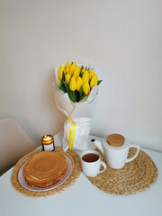 Still life with a bouquet of tulips, a teapot, a mug of tea, pancakes and a candle on a white table and kitchen. Spring table setting.