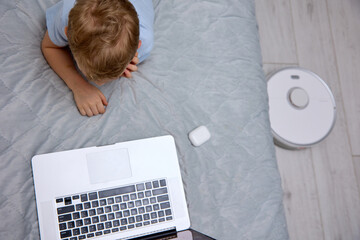 Little blond boy in white wireless headphones uses a modern laptop on the bed, gets new knowledge...