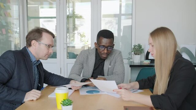 Ethnic diverse business team discuss new project sitting by table in office, meeting room. Creative group work together supporting each other. Corporate colleagues cooperating, analyzing graphs