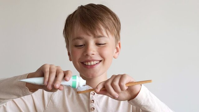 Child boy squeezes out white toothpaste onto eco toothbrush, smiling, isolated on light background. Boy's dental hygiene, morning brushing, lifestyle, medical care.