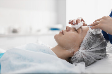 the cosmetologist washes off the cleansing foam from the face of the client with wet wipes before the main procedures