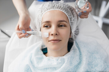 cosmetologist applies a moisturizing white cream mask to the patient's face with a brush