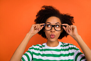Portrait of attractive minded funny girly wavy-haired girl touching specs learn pout lips isolated on bright orange color background