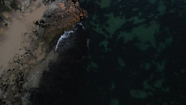 A bird's eye view footage of the open waters with various waves hitting the seashore. The aerial footage is rotating downward towards the water where you can see the rocks and green algae beneath.