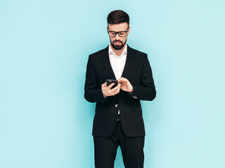 Handsome confident stylish hipster lambersexual model. Sexy modern man dressed in elegant black suit. Fashion male posing in studio. Holding smartphone. Looking at cellphone screen. Using apps