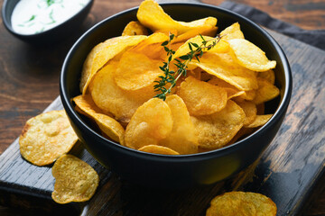 Potato chips. Fast food. Crispy potato chips ceramic black bowl with sour cream sauce and onions in wooden stand on old kitchen table wooden background. American tradition. Hot BBQ. Top view.