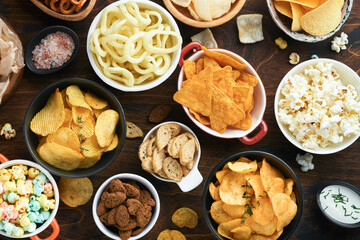 Unhealthy food or snacks. All classic potato snacks with peanuts, popcorn and onion rings and salted pretzels in bowl plates on old wooden background. Unhealthy food for figure, heart, skin, teeth.