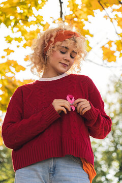 Vertical outdoor shot of a beautiful young woman suffering from breast cancer, reminding other people to do health checkups by holding pink ribbon. High quality photo
