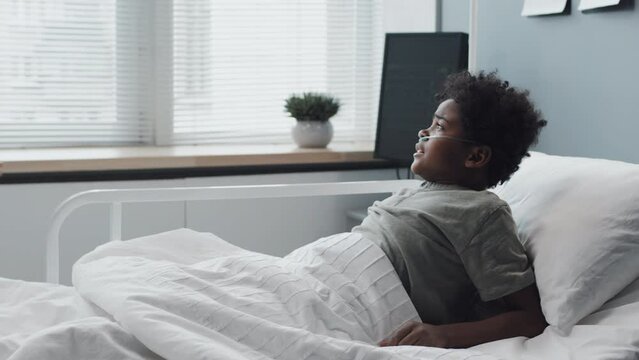 Close-up of African American boy with oxygen tube in nose sleeping in hospital bed at daytime, then waking up scared, mature Caucasian female doctor coming and comforting kid