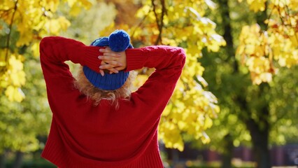 Laid-back caucasian young person standing backwards to the camera in red sweater and blue winter hat. Autumn colors trees in the background. High quality photo