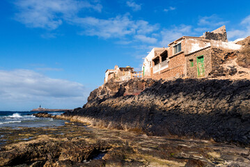 Vista de casas antiguas de Puerto de la Cruz que cuelgan en las rocas y el faro de Punta Jandia en el fondo, Fuerteventura, Islas Canarias, España