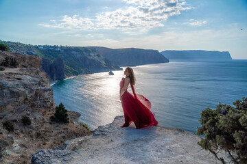 A girl with flowing hair in a long red dress stands on a rock above the sea. The stone can be seen in the sea. Sunny path to the sea from the sun.