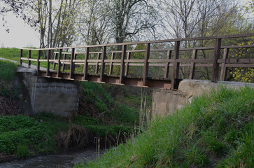 a park bridge with a wooden railing leads across the stream. The beams are fixed to a steel crossbeam that holds in a concrete foundation. road from gravel, in front of the bridge barrier