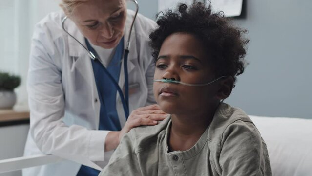 Tilting down and up of curly African American boy with oxygen tube sitting on bed in hospital room at daytime, blonde Caucasian female doctor using stethoscope on his back, talking to child