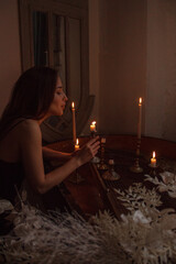 medieval night. young brunette slim girl in black silk dress stands in profile near grand piano with white flowers and blows out the candles on vintage room background. lifestyle concept, free space