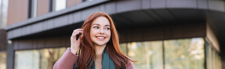 Fototapeta premium young pleased woman in scarf adjusting red hair near modern building, banner.