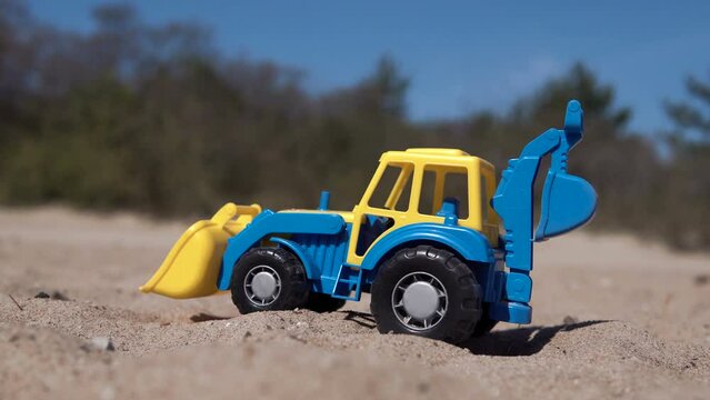 Little Boy plays with a tractor on the sand. Blue and yellow toy tractor. A surviving refugee from Ukraine plays on the beach. Sandbox with children's excavator. Picks up sand in a bucket. 4K
