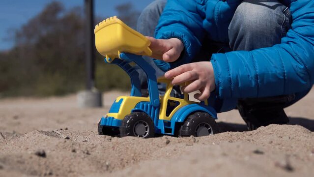 Little Boy plays with a tractor on the sand. Blue and yellow toy tractor. A surviving refugee from Ukraine plays on the beach. Sandbox with children's excavator. Picks up sand in a bucket. 4K